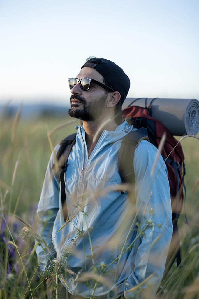 A man wearing sunglasses and carrying a backpack standing in a grassy field during a hike.
